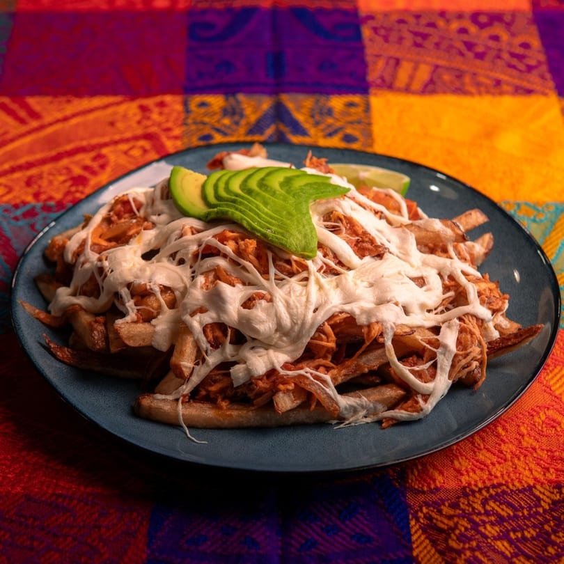 A plate of loaded fries with shredded chicken, melted cheese, and sliced avocado on a colorful tablecloth.