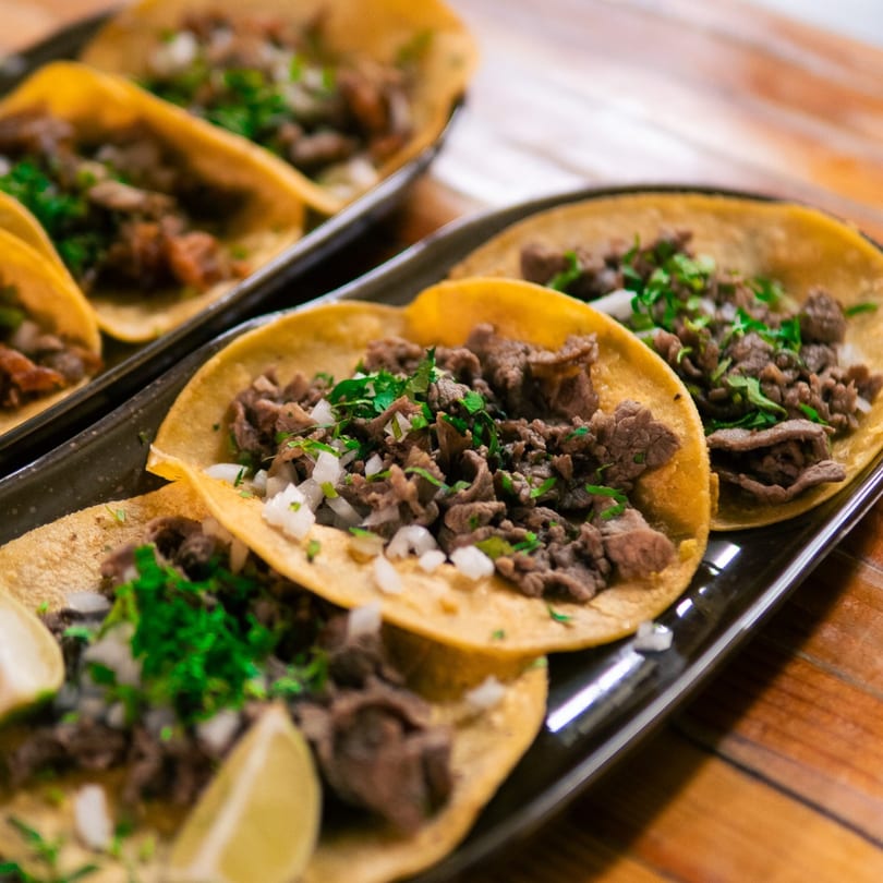 Close-up of several street-style tacos with meat, diced onions, and cilantro on a black plate.