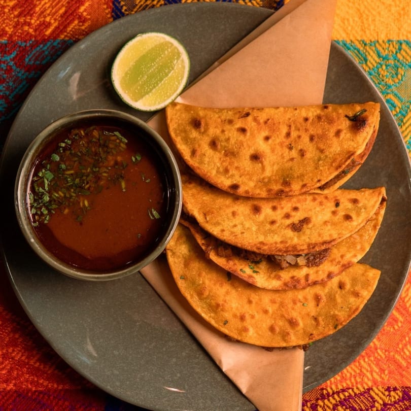 Four golden-brown birria tacos with a side of consommé and a lime wedge on a dark plate.